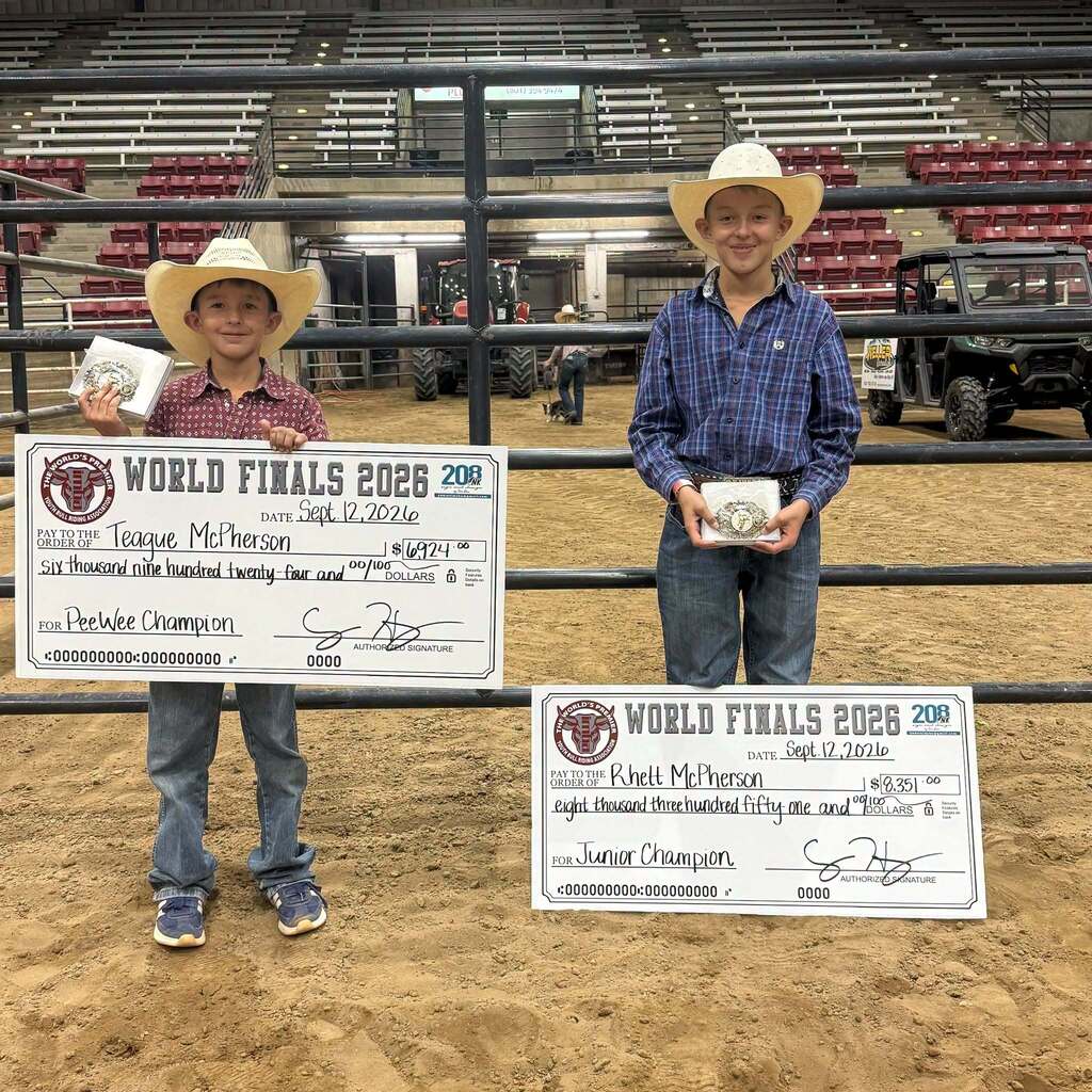 World Champions Teague and Rhett McPherson stand together in cowboy hats in the arena dirt, holding their championship belt buckles and large ceremonial checks from the World Finals.
