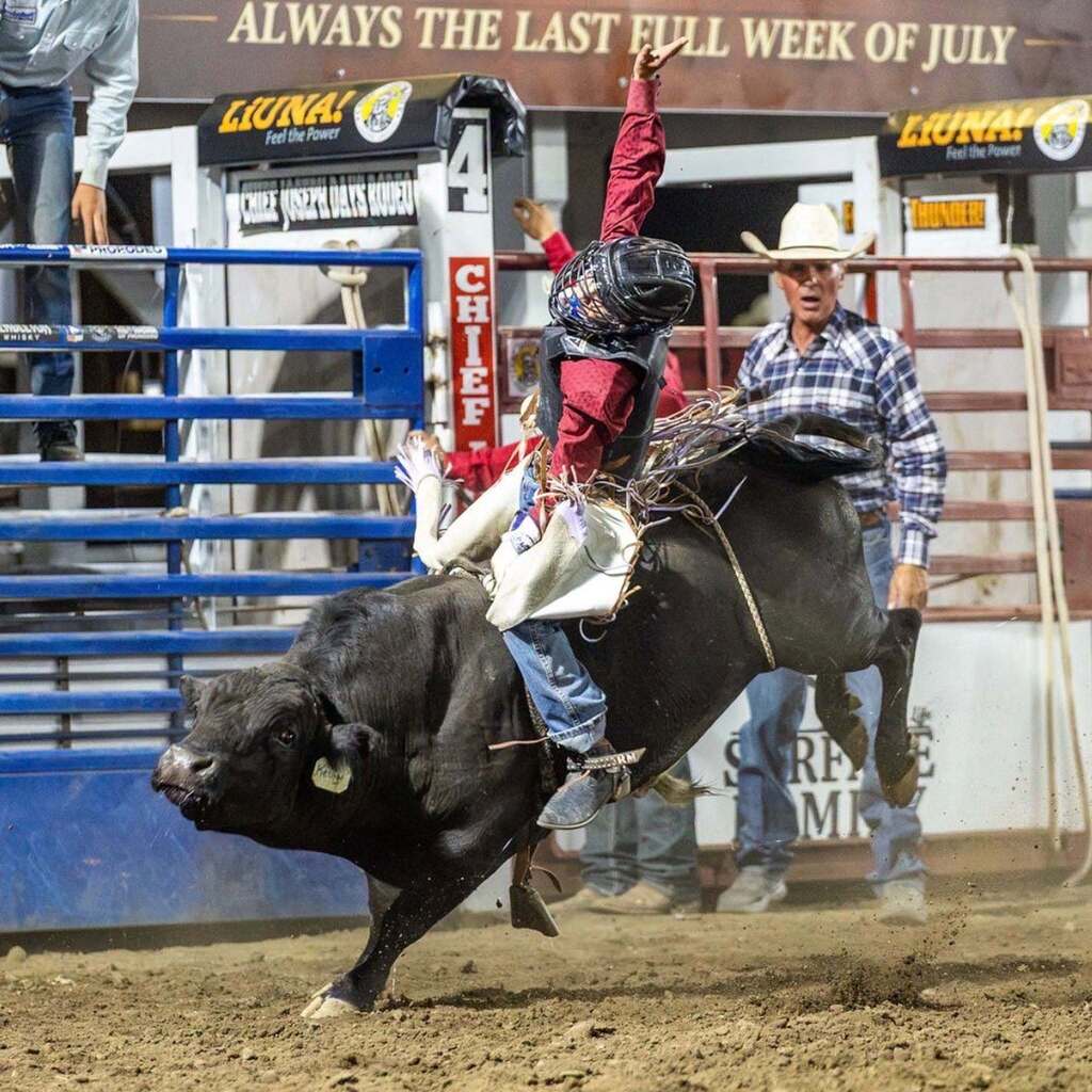 A young bull rider wearing a protective helmet and vest raises his free arm high while riding a bucking black bull out of the arena chutes.
