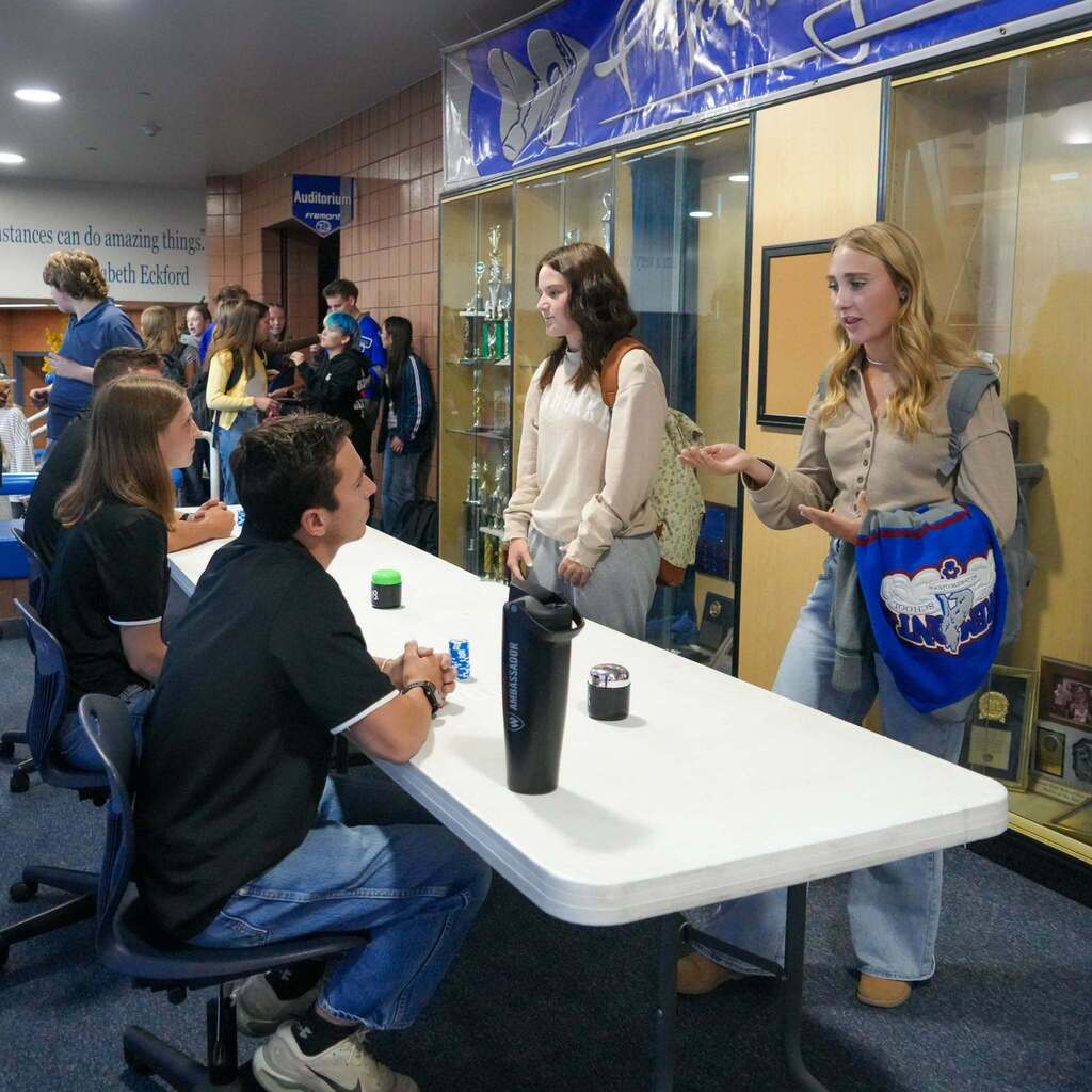 A Fremont High School student gestures animatedly with her hands while pitching a 60-second business solution to a guest judge seated across a white table in the school hallway.