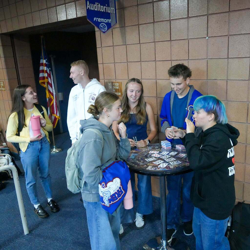 Students wearing school colors stand around a high-top table in the hallway, observing a card-based activity setup during the Wolf Tank event.