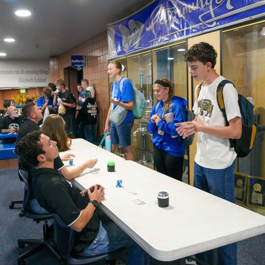 A student in a white graphic tee gestures with his hands while making a quick business pitch to a judge seated at a long table in the busy hallway.