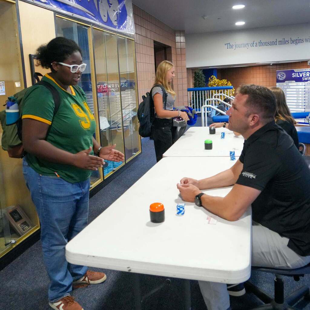 A female student wearing a green and gold shirt uses expressive hand gestures to pitch her quick business ideas to a guest judge from Weber State University.
