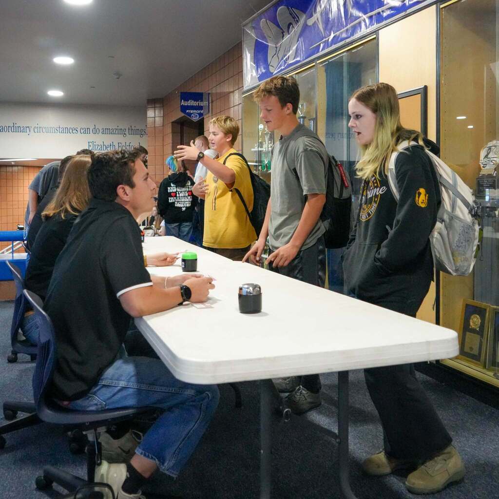 Two male high school students present their pitch to a judge seated at a white table, with school award trophy cases visible in the background.