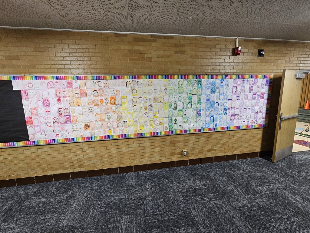 A wide view of a long bulletin board in a school hallway filled with hundreds of student self-portraits arranged in a continuous color gradient from red to purple.