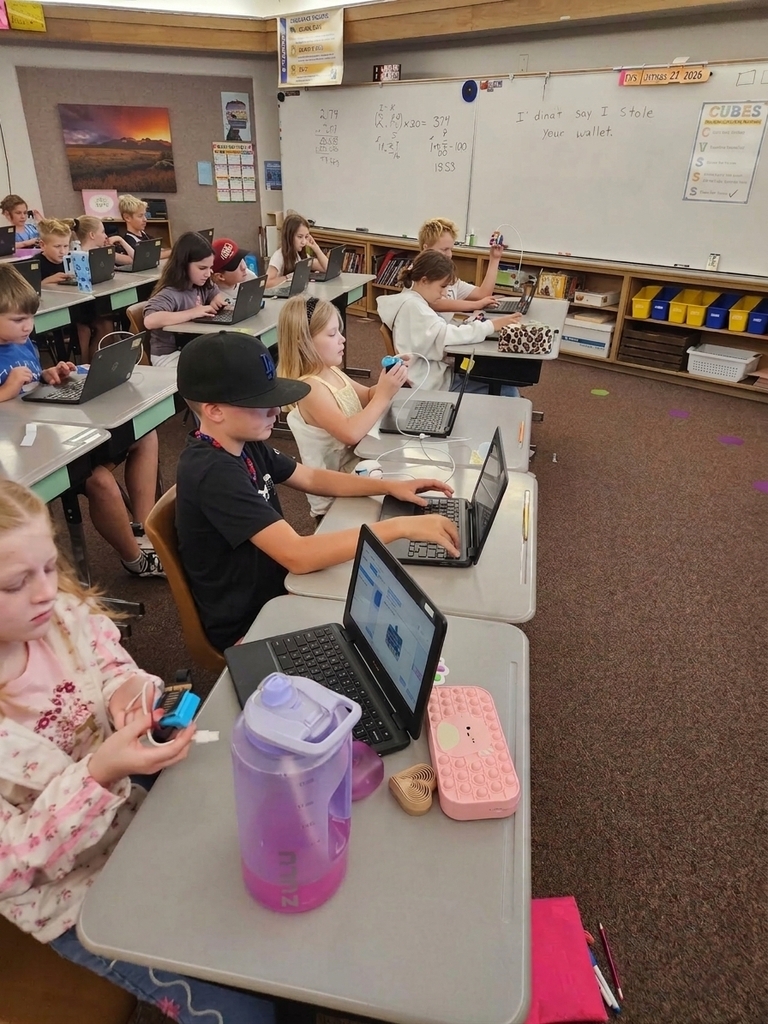 Students sitting at classroom desks using laptops and handling micro:bit programmable microcontrollers with battery packs during a coding lesson.