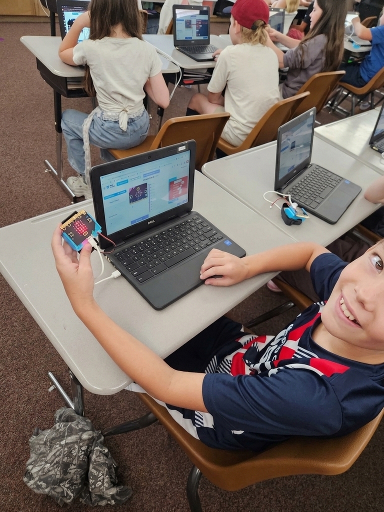A smiling student at a desk holding up a micro:bit device connected to a laptop running a block-based coding program.