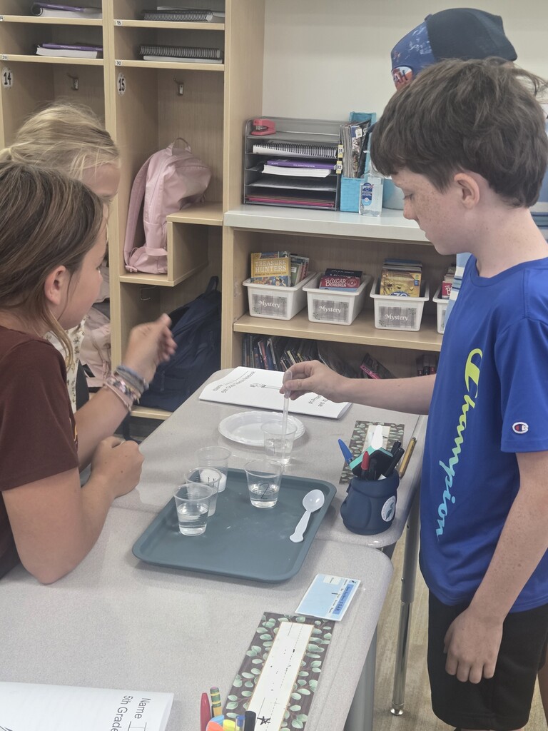 A boy in a blue Champion shirt uses a pipette at a lab station with cups of liquid while classmates watch. Notebooks on the desk read "5th Grade Science Notebook" and "#1-Properties and #2- Models", with cubbies behind labeled "Mystery".