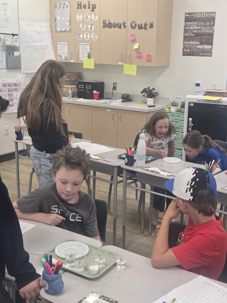 Students work on a science lab at grouped desks, featuring a boy in a grey "SPACE" shirt near liquid sample cups. A cart in the room is labeled "143-CART-B204", and wall posters read "Help", "Shout Outs", and "TODAY"