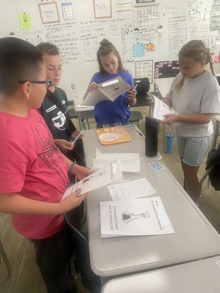 Four students compare their "5th Grade Science Notebook" packets around an experiment setup. Wall posters in the background show "Language Arts", "Math", and calendar notes for "SEPTEMBER"