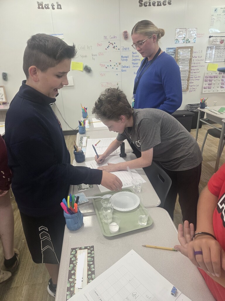 A teacher in a blue cardigan assists students as they conduct a liquid science experiment. Whiteboards behind them display "Math", "Science", "IXL Codes", and "Zearn".