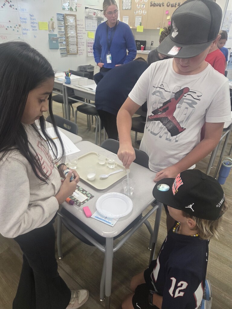 A student in a Jordan shirt uses a pipette over liquid cups while group members observe. Wall signs in the background display "Help", "Shout Outs", and handwritten lesson notes.
