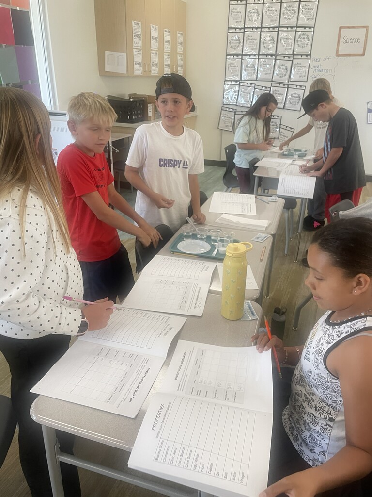 A student fills out a worksheet titled "MYSTERY LIQUID INVESTIGATION" listing items like "Baking Soda", "Cornstarch", "Citric Acid", "Flour", "Sugar", and "Vinegar". A boy in a shirt reading "CRISPY" works nearby.