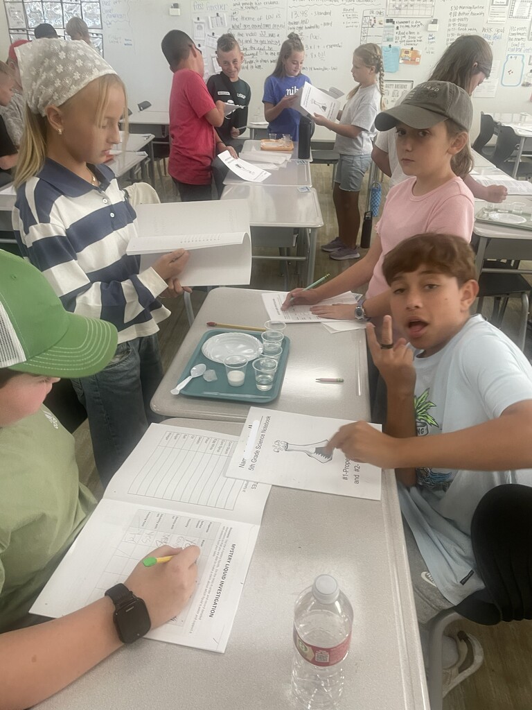 A student fills out a worksheet titled "MYSTERY LIQUID INVESTIGATION" listing items like "Baking Soda", "Cornstarch", "Citric Acid", "Flour", "Sugar", and "Vinegar". A boy in a shirt reading "CRISPY" works nearby.