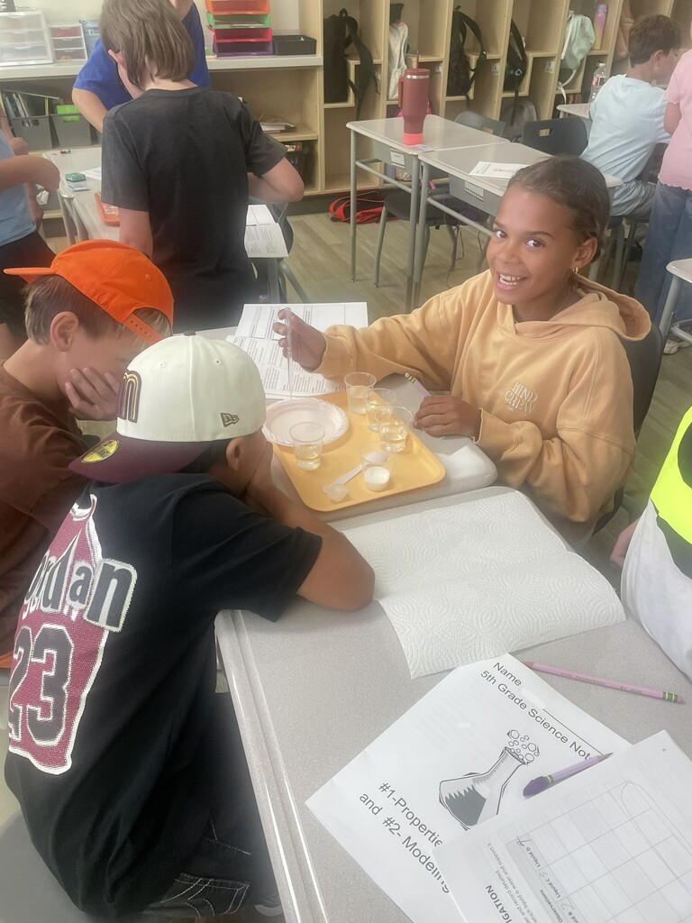 A smiling girl in a yellow hoodie sits at a science lab station with liquid samples. A booklet on the desk reads "5th Grade Science Notebook", "#1-Properties and #2- Models".