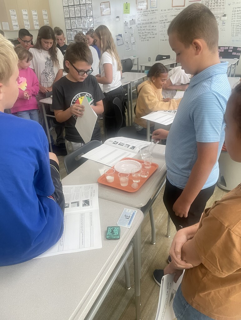 Students gather around a tray of liquid samples, with one holding a packet and another in a black shirt reading "LOVE". Wall displays behind them feature "Language Arts", "Math", "SEPTEMBER", and lesson objectives.