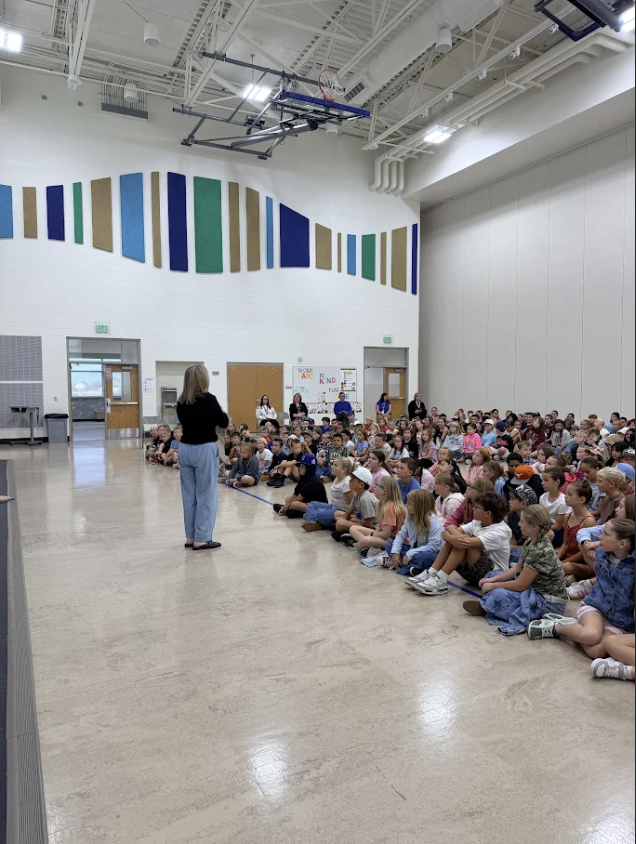 Side perspective of a school gymnasium where a speaker in a black top and light pants addresses a large group of students seated on the floor along the wall. In the background, a white banner on the wall reads "BE KIND".