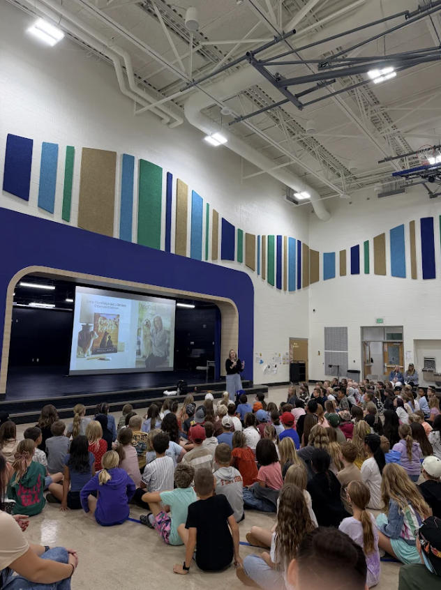 High-angle shot of a full school gymnasium filled with students sitting on the floor watching an author presentation. The stage screen displays the book cover for Forever Ripley alongside the text "McCall Hoyle"
