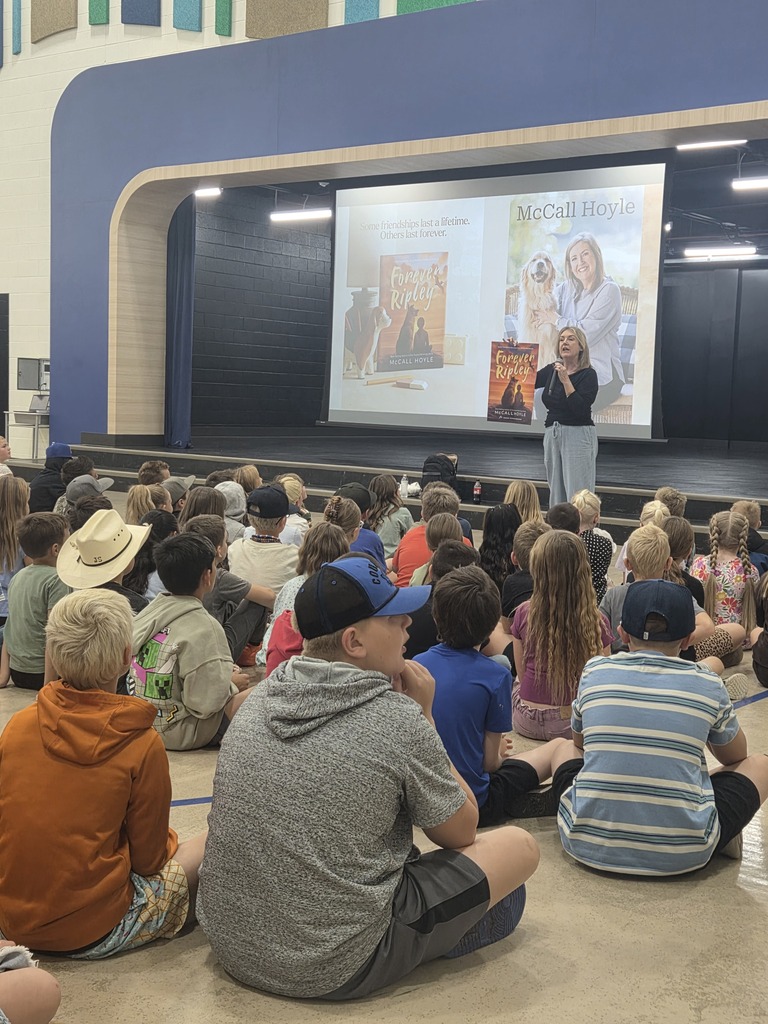 Author McCall Hoyle stands on a stage presenting to a large audience of elementary students seated on the floor. On the projection screen behind her, text reads "Some friendships last a lifetime. Others last forever.", "Forever Ripley", "McCall Hoyle", and "McCall Hoyle". She holds a copy of her book Forever Ripley.