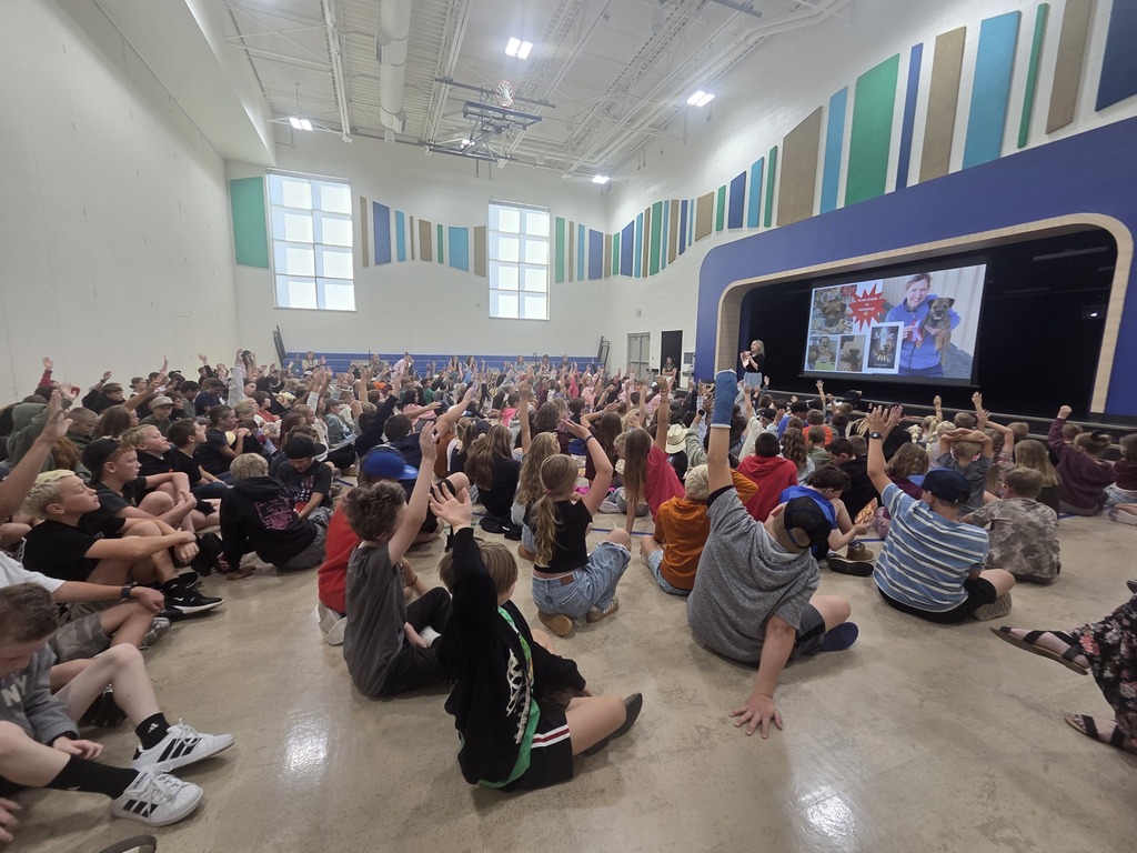 Wide view of a school auditorium filled with seated students raising their hands during a presentation. A projection screen on stage displays images of a woman with dogs, along with book cover art and text that includes "Even Edith is amazing!" and "Millie".