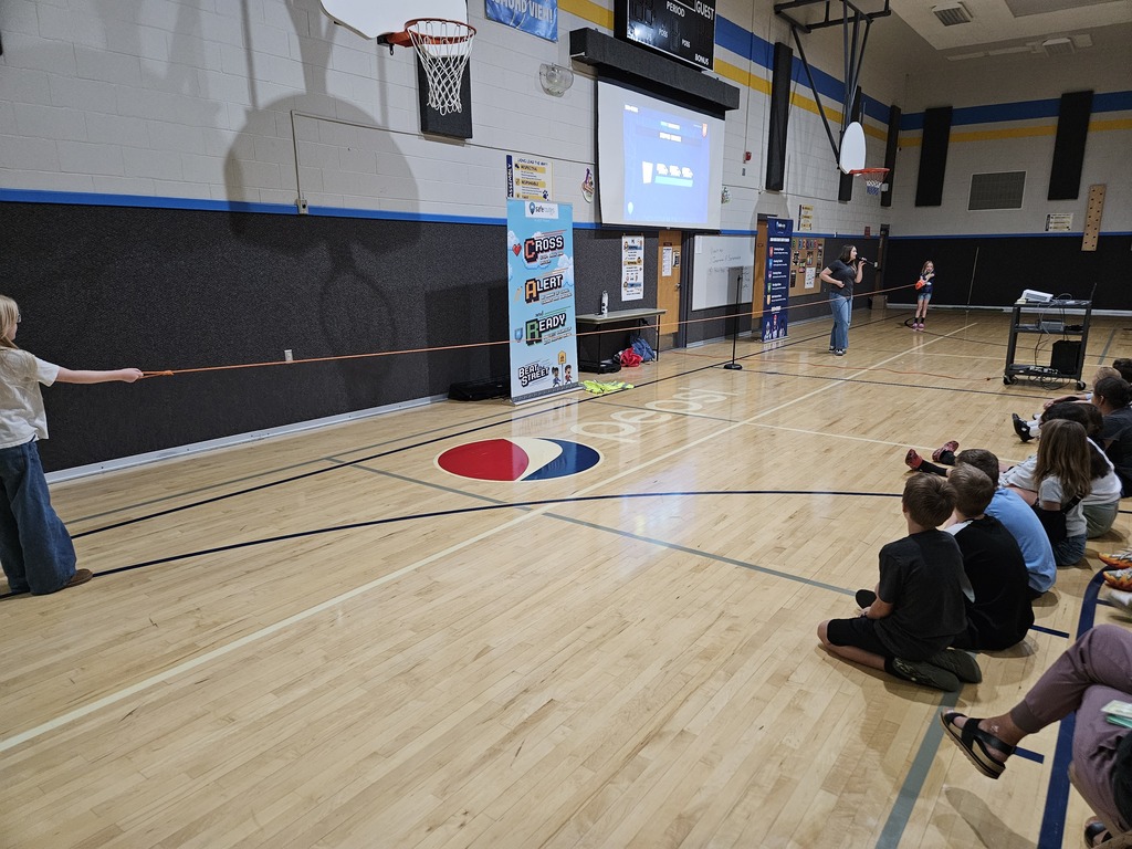 Students sitting on the gymnasium floor watching a Safe Routes Utah assembly demonstration where a long orange measuring tape shows how far a car takes to stop.