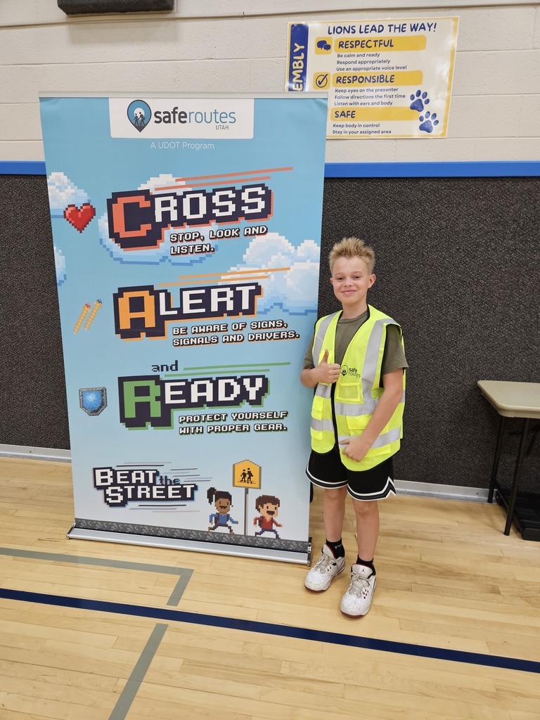 A student giving a thumbs-up next to a street safety banner while wearing a high-visibility neon yellow safety vest.