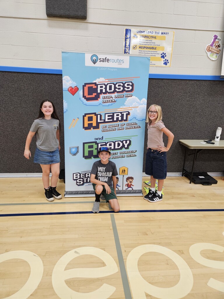 Three students posing in front of a "Safe Routes Utah - Cross, Alert, and Ready" banner in the gym.