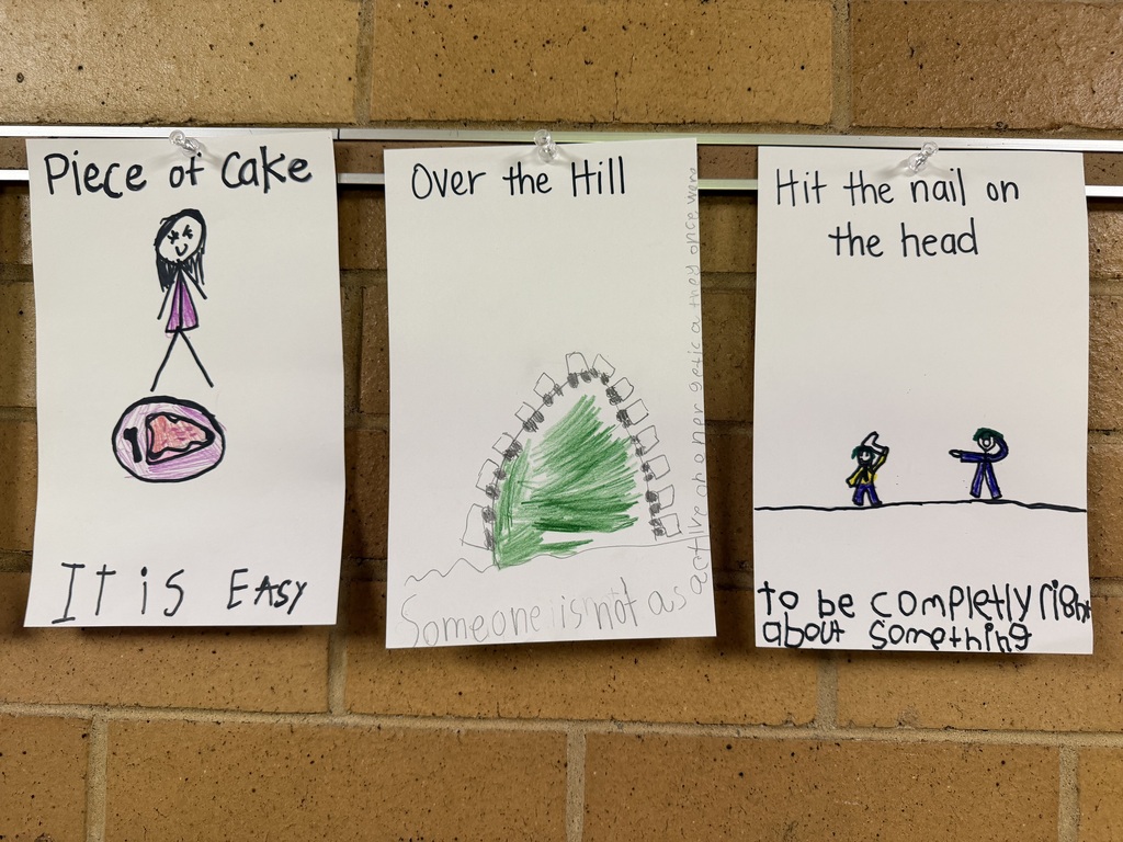 Student drawings displayed on a wall illustrating the idioms "Piece of Cake," "Over the Hill," and "Hit the nail on the head."