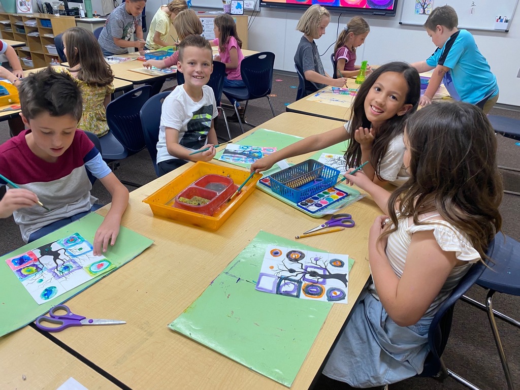 students smiling while sitting at a long table with paint brushes in hand and a colorful page before them with spooky trees painted over colorful shapes