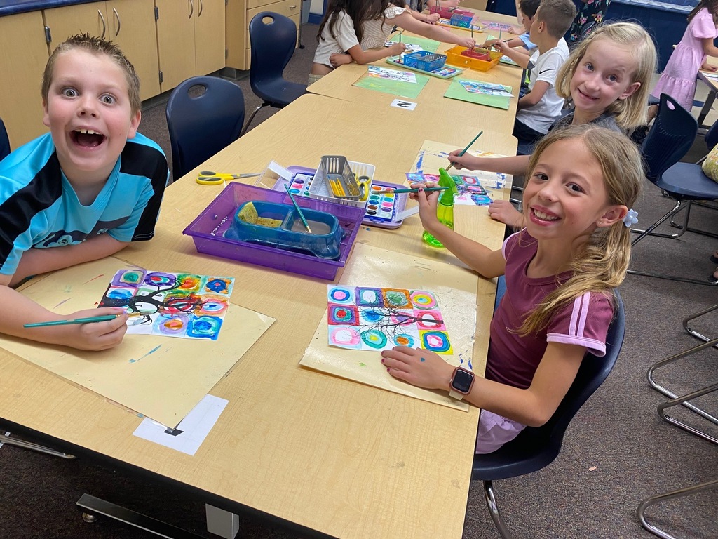 students smiling while sitting at a long table with paint brushes in hand and a colorful page before them with spooky trees painted over colorful shapes