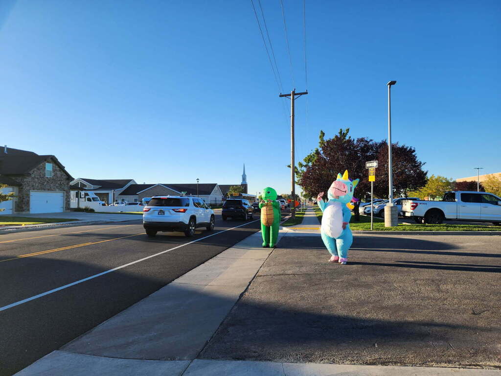 Two school staff members wearing inflatable costumes—a green turtle and a light blue unicorn with a pink mane and yellow horn—stand on a sunny sidewalk waving to passing cars outside a school. A white SUV and other vehicles line the two-lane road next to them, with suburban houses and a church steeple in the background under a clear blue sky.