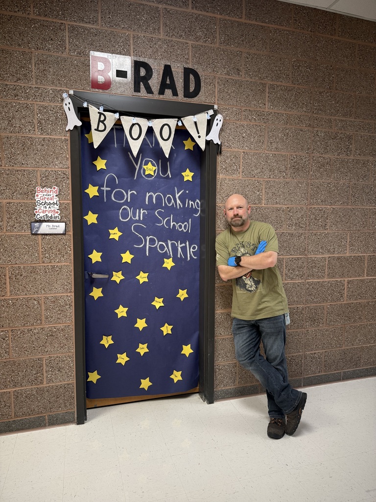 A man stands beside a decorated school doorway. Above the doorway is a sign reading “B-RAD,” and a Halloween-themed banner reads “BOO!” with ghost cutouts. The door is covered in dark blue paper with yellow stars and a handwritten message thanking him for “making our school sparkle.” A small sign identifies him as Mr. Brad, the custodian. He is wearing a green T-shirt, jeans, and work boots, with his arms crossed and one leg crossed in front of the other.