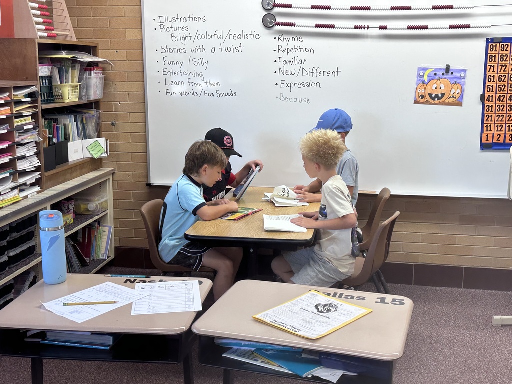 Four young students sitting together around a small table in front of a whiteboard reading books together. 