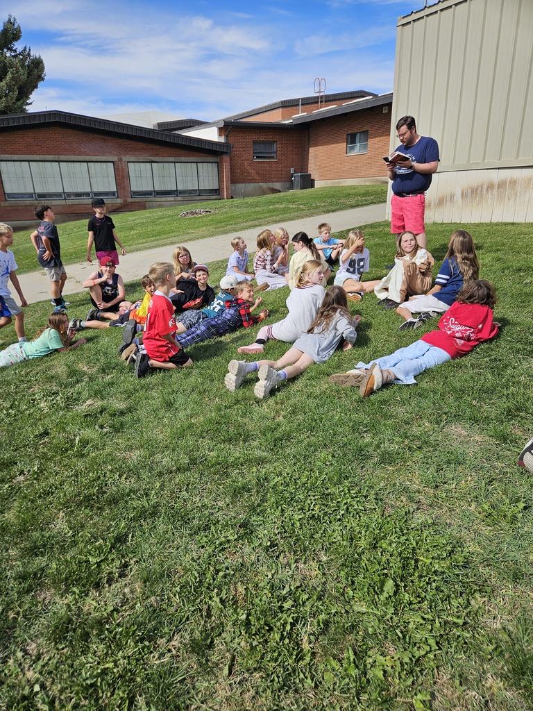 A class sitting outside on the grass under a blue sky listening to a teacher read aloud from a book.