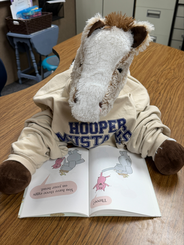 A stuffed horse is seeing wearing a shirt that says Hooper Mustangs and is reading a picture book