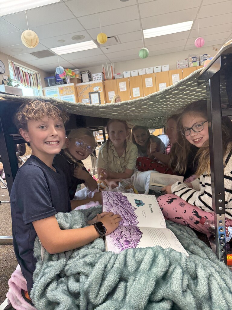 A group of elementary students sits together under a blanket fort, reading books and smiling for the camera in their classroom.