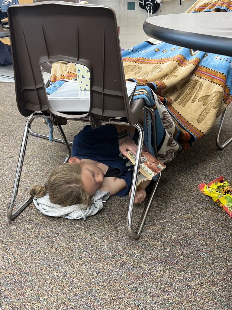 A student lies on the classroom floor beneath a blanket-covered desk, reading a book while surrounded by blankets, pillows, and snacks.