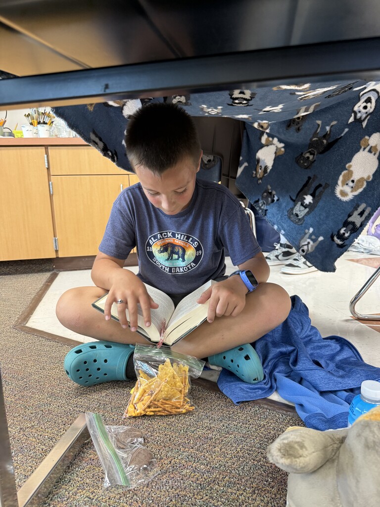 An elementary student sits cross-legged beneath a blanket-covered desk, reading a book with a snack bag nearby.