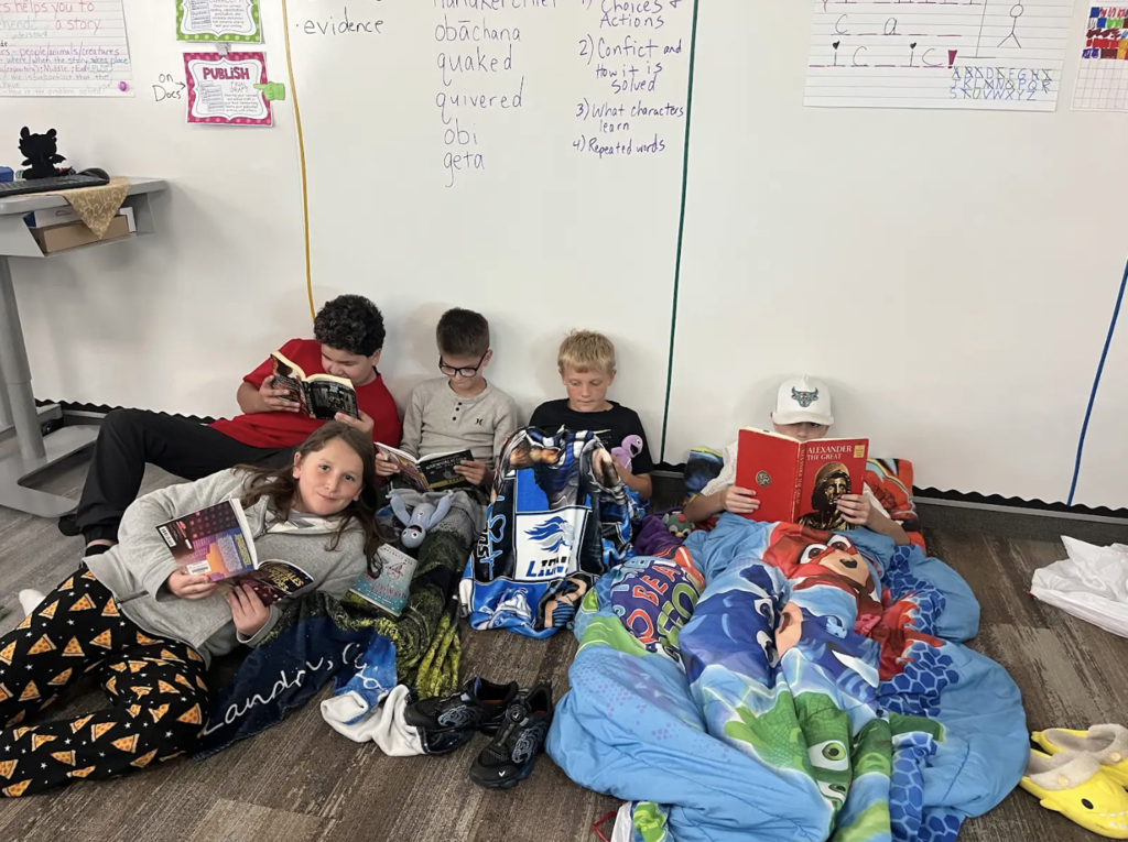 5 boys leaning against a white board reading books while laying with blankets
