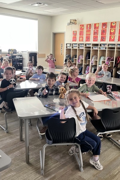 Elementary students sitting at desks in a classroom holding small flashlights, stuffed animals, and books. Visible text includes phonics posters on the cubbies (such as "sh", "th", "ch", "qu") and "HAVEN" on a girl's white t-shirt.