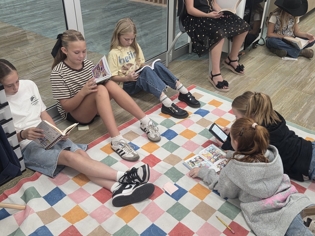 Students sitting and laying on a colorful checkered rug on the floor reading books and a digital device. Visible text includes "hello" on a yellow t-shirt and "OFF THE WALL" on a white t-shirt.