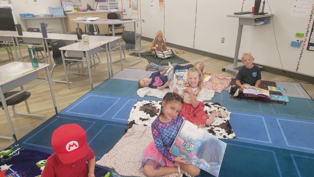 Children sitting on blankets on a blue tiled classroom carpet reading picture books, with one boy wearing a red Mario hat. Visible text includes "M" on the red hat, "YOSHI" on a blanket, and "B" on a wall outlet plate.