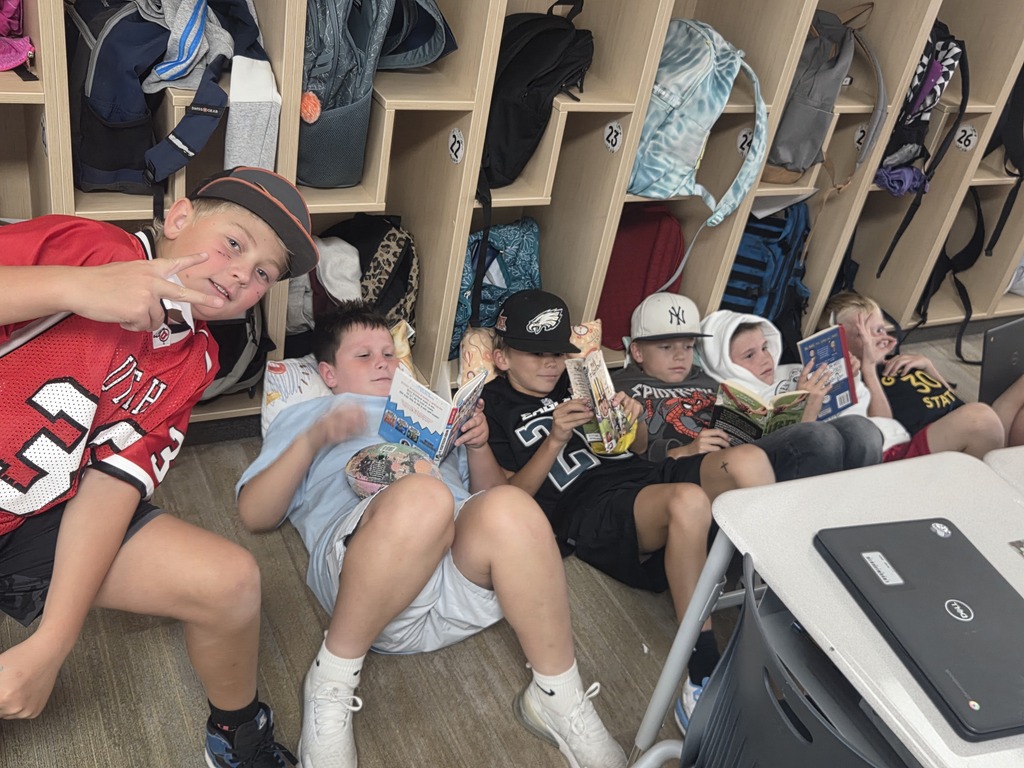 A group of young boys sitting on the floor leaning against cubbies while reading books, with one boy giving a peace sign. Visible text includes "33" on a red jersey, "22", "23", "24", "25", "26" on cubbies, "SPIDER-MAN" on a shirt, "GOAT 30 STAT" on a shirt, and "DELL" on a laptop screen.