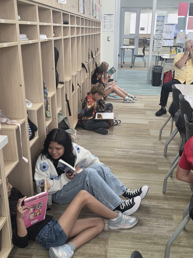 Students sitting on the floor along a hallway lined with cubbies reading books, with a teacher sitting at a desk nearby. Visible text includes "IN AN EMERGENCY" on a wall poster and "CUPCAKE DIARIES" on a pink book cover.