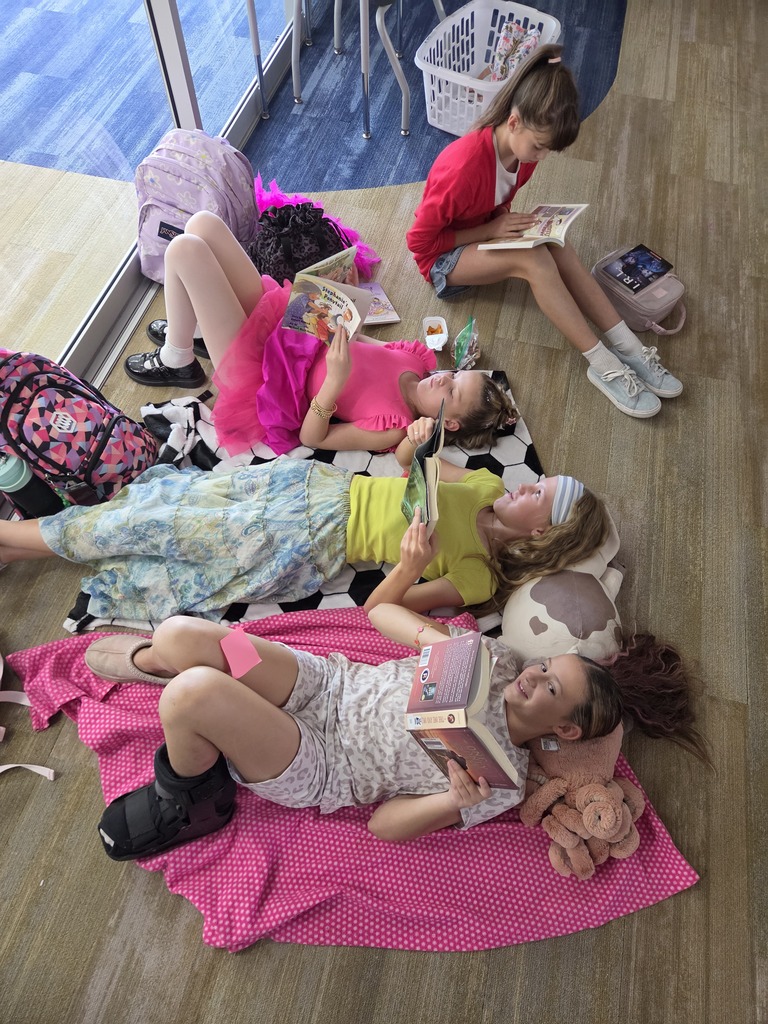 Four young girls laying down on blankets on a classroom floor while reading books. Visible text includes "Stephanie's Ponytail" on a book cover and "I.R.L." on a pencil pouch.