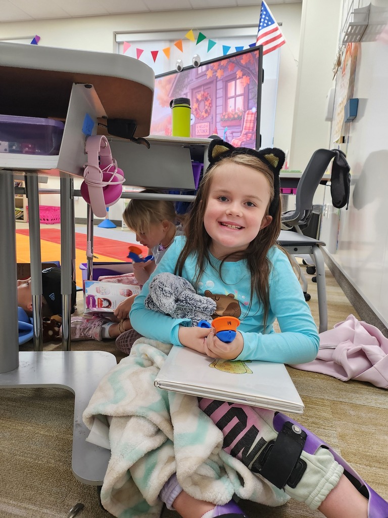 A young girl wearing cat ears and holding a plush stuffed animal smiles while sitting on the floor under a desk with a book in her lap. Visible text includes "OCTOBER" on a monitor screen, "Glossary" on a book spine, "19" on a orange tool, and "PINK" on her leggings.