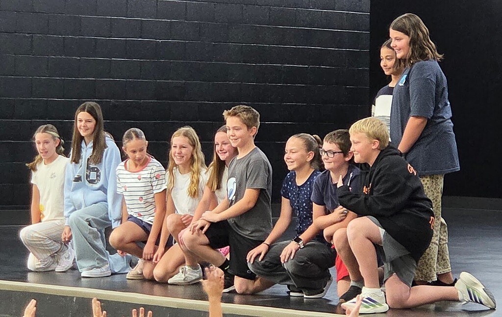 A line of ten elementary students kneeling and standing on a stage during a presentation, with a dark brick wall in the background.