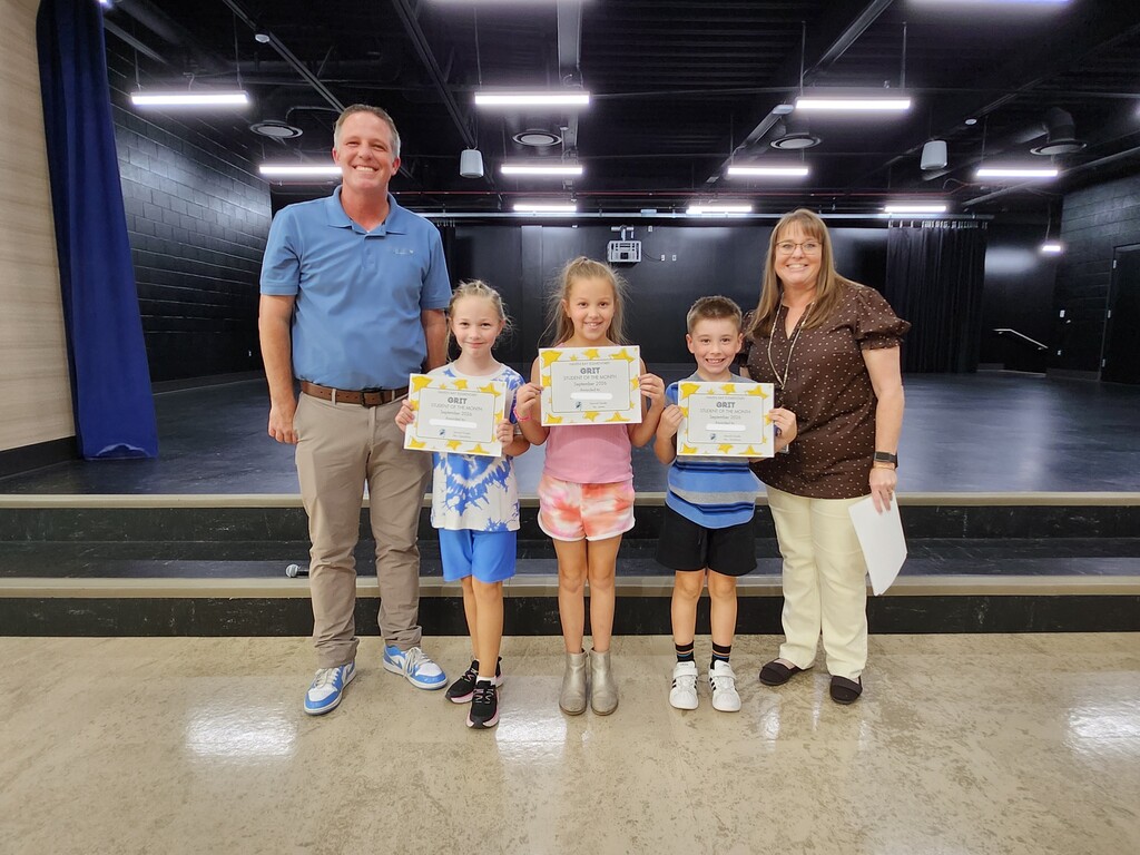 Mr. Skeen and a Mrs. Widdison standing alongside three second grade students holding "GRIT - Student of the Month - September 2024" certificates on stage.