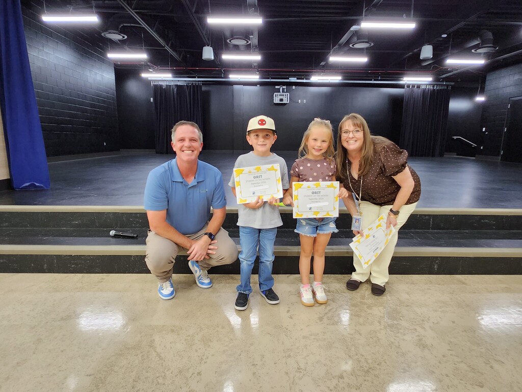 Mr. Skeen crouching and a Mrs. Widdison standing alongside two young students holding "GRIT - Student of the Month - September 2024" certificates on stage.