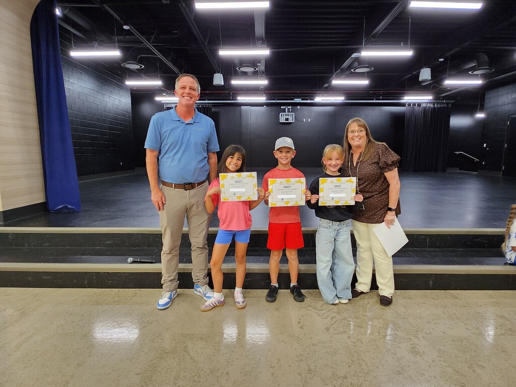 Mr. Skeen and a Mrs. Widdison standing alongside three fourth grade students holding "GRIT - Student of the Month - September 2024" certificates on stage.