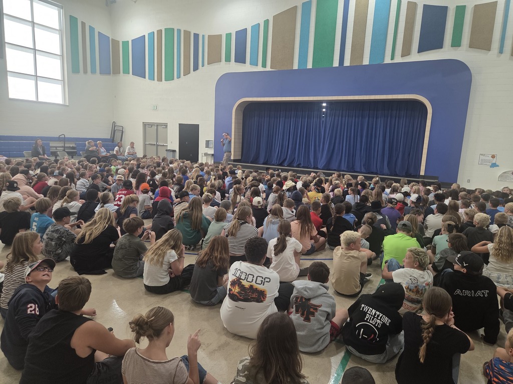 A large group of elementary students sitting on the floor facing a stage during a school assembly in a brightly lit auditorium with colourful wall paneling.